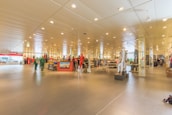 A spacious, well-lit retail store interior featuring various clothing displays and mannequins. The lighting is bright with numerous ceiling lights illuminating the area. Clothing racks and sections can be seen throughout the space, with colorful apparel on display. The store has a modern design with tiled flooring and a systematic arrangement of merchandise.
