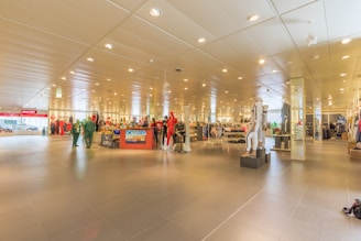 A spacious, well-lit retail store interior featuring various clothing displays and mannequins. The lighting is bright with numerous ceiling lights illuminating the area. Clothing racks and sections can be seen throughout the space, with colorful apparel on display. The store has a modern design with tiled flooring and a systematic arrangement of merchandise.