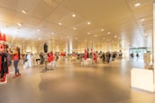 A spacious, well-lit clothing store features neatly organized racks of clothes and accessories. Several mannequins display stylish outfits, and a few customers are browsing through the aisles. The interior is modern with a high ceiling adorned with numerous lights that create a bright and inviting atmosphere.