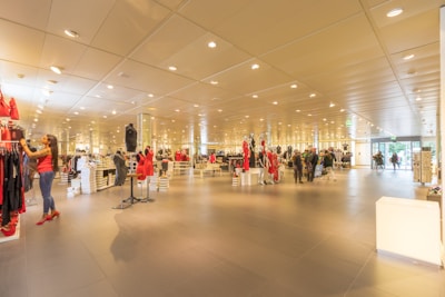 A spacious, well-lit clothing store features neatly organized racks of clothes and accessories. Several mannequins display stylish outfits, and a few customers are browsing through the aisles. The interior is modern with a high ceiling adorned with numerous lights that create a bright and inviting atmosphere.