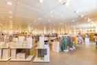 A brightly lit, spacious retail clothing store with rows of neatly arranged clothing racks and shelves filled with folded garments. The store has high ceilings with numerous spotlights and a modern, minimalist design. Multiple stacks of white shoe boxes are prominently displayed on tables throughout the space.