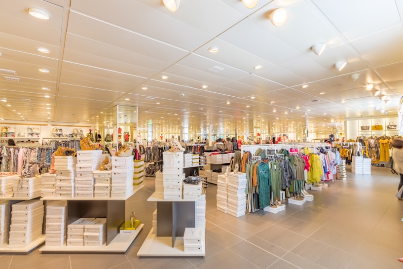 A brightly lit, spacious retail clothing store with rows of neatly arranged clothing racks and shelves filled with folded garments. The store has high ceilings with numerous spotlights and a modern, minimalist design. Multiple stacks of white shoe boxes are prominently displayed on tables throughout the space.