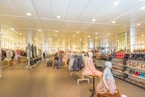 A spacious retail store with bright lighting and neatly arranged clothing and footwear displays. Mannequins wearing children's outfits are prominently featured in the foreground, while various clothes and shoes are organized on racks and shelves throughout the store. People are seen browsing and shopping in the background, adding a lively atmosphere to the setting.