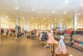 A spacious retail store with bright lighting and neatly arranged clothing and footwear displays. Mannequins wearing children's outfits are prominently featured in the foreground, while various clothes and shoes are organized on racks and shelves throughout the store. People are seen browsing and shopping in the background, adding a lively atmosphere to the setting.