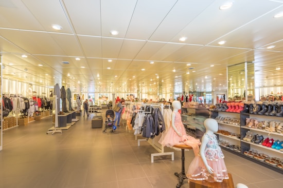 A spacious retail store with bright lighting and neatly arranged clothing and footwear displays. Mannequins wearing children's outfits are prominently featured in the foreground, while various clothes and shoes are organized on racks and shelves throughout the store. People are seen browsing and shopping in the background, adding a lively atmosphere to the setting.