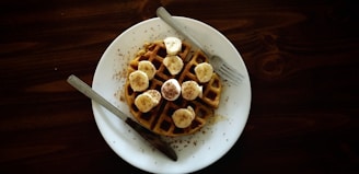 waffle with sliced banana fruits on top near gray stainless steel fork and knife
