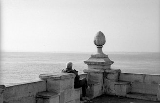 An older person with short, light-colored hair sits on a stone terrace overlooking a calm sea. The terrace features classic stone balustrades and a notable decorative stone ornament. The scene exudes a sense of tranquility and introspection.