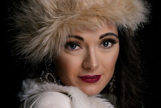 A close-up portrait of a person with striking facial features, wearing a fluffy, cream-colored hat and a matching fur-collared coat. The makeup is bold, with dark, well-defined eyebrows, long eyelashes, and deep red lipstick. The person's expression is confident and direct, with earrings visible that add a touch of elegance.