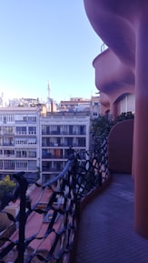 A uniquely designed balcony with an undulating, organic form overlooks a cityscape. The balcony features an artistic black wrought iron railing and is lined with greenery. In the background, a series of modern apartment buildings are visible under a clear blue sky.