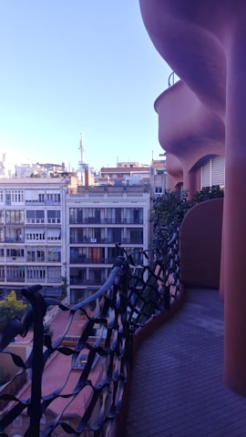 A uniquely designed balcony with an undulating, organic form overlooks a cityscape. The balcony features an artistic black wrought iron railing and is lined with greenery. In the background, a series of modern apartment buildings are visible under a clear blue sky.