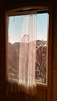Ornate sheer curtains with intricate floral patterns hang on a window, filtering the warm sunlight. The view outside shows urban buildings under a clear blue sky and architectural elements of a balcony railing.