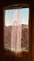 Ornate sheer curtains with intricate floral patterns hang on a window, filtering the warm sunlight. The view outside shows urban buildings under a clear blue sky and architectural elements of a balcony railing.