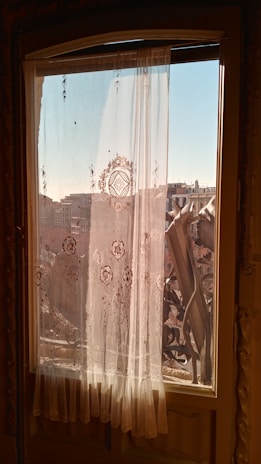Ornate sheer curtains with intricate floral patterns hang on a window, filtering the warm sunlight. The view outside shows urban buildings under a clear blue sky and architectural elements of a balcony railing.