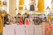 A well-lit clothing and accessories store showcases high heel shoes in various colors, arranged on top of white boxes. Behind the shoes, there are racks and shelves displaying yellow and mustard dresses, handbags, and a few mannequins wearing stylish outfits.