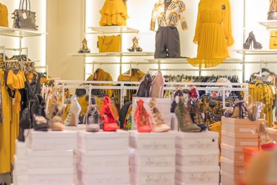 A well-lit clothing and accessories store showcases high heel shoes in various colors, arranged on top of white boxes. Behind the shoes, there are racks and shelves displaying yellow and mustard dresses, handbags, and a few mannequins wearing stylish outfits.