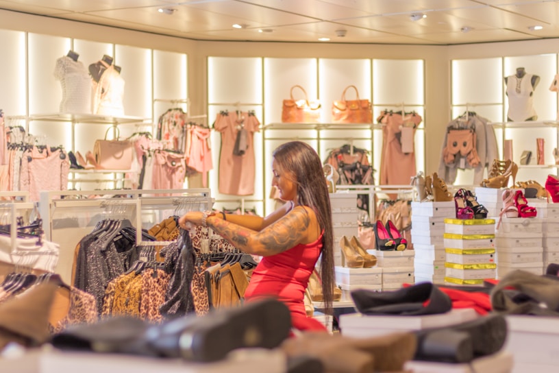 A woman with long hair and tattoos browses through racks of clothing, surrounded by displays of colorful shoes, handbags, and dresses in a well-lit fashion store. The atmosphere is vibrant and stylish, showcasing a variety of fashionable items.
