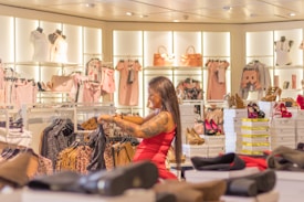 A woman with long hair and tattoos browses through racks of clothing, surrounded by displays of colorful shoes, handbags, and dresses in a well-lit fashion store. The atmosphere is vibrant and stylish, showcasing a variety of fashionable items.