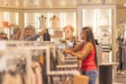 Two women are shopping in a brightly lit clothing store. They are browsing through racks of clothes, with various apparel and accessories displayed on shelves and hangers in the background. One woman is holding up a garment to show the other.