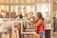 Two women are shopping in a brightly lit clothing store. They are browsing through racks of clothes, with various apparel and accessories displayed on shelves and hangers in the background. One woman is holding up a garment to show the other.