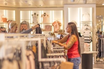 Two women are shopping in a brightly lit clothing store. They are browsing through racks of clothes, with various apparel and accessories displayed on shelves and hangers in the background. One woman is holding up a garment to show the other.