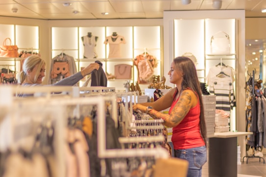 Two women are shopping in a brightly lit clothing store. They are browsing through racks of clothes, with various apparel and accessories displayed on shelves and hangers in the background. One woman is holding up a garment to show the other.