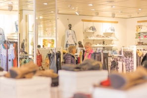 A clothing store interior with racks of colorful garments and accessories. Two people are browsing through clothing. The store is well-lit, with mannequins displaying outfits and shelves stocked with bags.