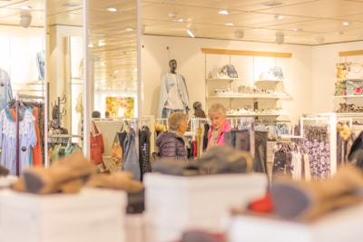 A clothing store interior with racks of colorful garments and accessories. Two people are browsing through clothing. The store is well-lit, with mannequins displaying outfits and shelves stocked with bags.