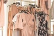 A collection of dresses and clothing items hanging on a rack. The garments include a variety of pastel colors and floral patterns, with predominant hues of blush pink. The clothing is displayed in a well-organized manner, suggesting a fashion store or boutique setting.