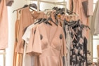 A collection of dresses and clothing items hanging on a rack. The garments include a variety of pastel colors and floral patterns, with predominant hues of blush pink. The clothing is displayed in a well-organized manner, suggesting a fashion store or boutique setting.