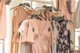 A collection of dresses and clothing items hanging on a rack. The garments include a variety of pastel colors and floral patterns, with predominant hues of blush pink. The clothing is displayed in a well-organized manner, suggesting a fashion store or boutique setting.