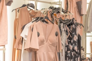 A collection of dresses and clothing items hanging on a rack. The garments include a variety of pastel colors and floral patterns, with predominant hues of blush pink. The clothing is displayed in a well-organized manner, suggesting a fashion store or boutique setting.