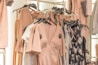 A collection of dresses and clothing items hanging on a rack. The garments include a variety of pastel colors and floral patterns, with predominant hues of blush pink. The clothing is displayed in a well-organized manner, suggesting a fashion store or boutique setting.