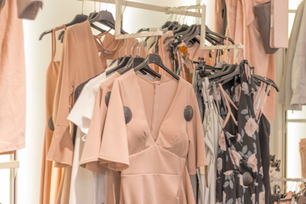 A collection of dresses and clothing items hanging on a rack. The garments include a variety of pastel colors and floral patterns, with predominant hues of blush pink. The clothing is displayed in a well-organized manner, suggesting a fashion store or boutique setting.