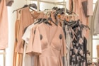 A collection of dresses and clothing items hanging on a rack. The garments include a variety of pastel colors and floral patterns, with predominant hues of blush pink. The clothing is displayed in a well-organized manner, suggesting a fashion store or boutique setting.