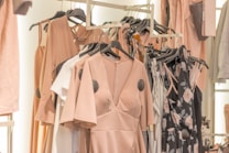 A collection of dresses and clothing items hanging on a rack. The garments include a variety of pastel colors and floral patterns, with predominant hues of blush pink. The clothing is displayed in a well-organized manner, suggesting a fashion store or boutique setting.