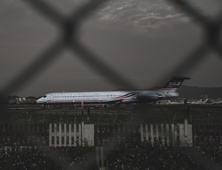 A commercial airplane is parked on a runway, with a focus on the side view through a wire fence. The airplane has the logo of Far Eastern Air Transport, and is set against the backdrop of a cloudy sky and distant mountains. A picket fence and grass are visible in the foreground.