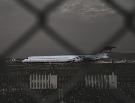 A commercial airplane is parked on a runway, with a focus on the side view through a wire fence. The airplane has the logo of Far Eastern Air Transport, and is set against the backdrop of a cloudy sky and distant mountains. A picket fence and grass are visible in the foreground.