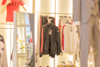 A retail store interior displaying various clothing items, including jackets and sweaters on racks. Mannequins dressed in outfits, one wearing a red shirt. Shoes are visible on the shelves in the background.