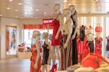 Mannequins are displayed in a brightly lit clothing store, adorned in stylish dresses. The background features shelves with various red handbags and shoes. Art can be seen on the walls, adding a decorative touch.