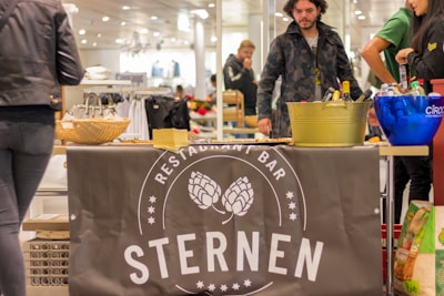 A market stall with a banner featuring the text 'Restaurant Bar Sternen' displayed. Various beverages are organized in ice buckets including a blue Ciroc bucket, and there are snacks on the table, possibly samples. People are standing around the stall in casual clothing.