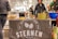 A market stall with a banner featuring the text 'Restaurant Bar Sternen' displayed. Various beverages are organized in ice buckets including a blue Ciroc bucket, and there are snacks on the table, possibly samples. People are standing around the stall in casual clothing.