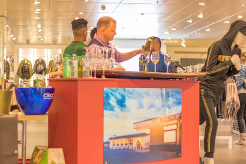 A group of people gathered around a vendor's stand inside a store. The stand is decorated with a poster displaying an image of a building labeled 'Weinkellerei Noesch'. Various bottles and glasses are displayed, suggesting a wine or beverage tasting event. A mannequin dressed in black sportswear is visible nearby, indicating a retail environment.