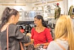 Three women are gathered in a fashion store setting, smiling and engaging in conversation. The background features shelves with clothing and a sign that reads 'Courage Mode'. Bags with red hearts are visible behind them.