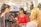 Three women are gathered in a fashion store setting, smiling and engaging in conversation. The background features shelves with clothing and a sign that reads 'Courage Mode'. Bags with red hearts are visible behind them.