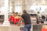 Interior of a clothing store featuring a man interacting with two women behind the counter. The store appears to be well-lit with chandeliers and has various clothing items displayed in the background.