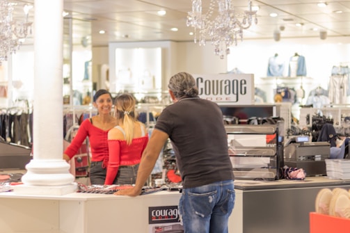Interior of a clothing store featuring a man interacting with two women behind the counter. The store appears to be well-lit with chandeliers and has various clothing items displayed in the background.