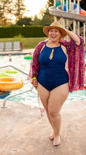 A person wearing a blue one-piece swimsuit and a wide-brimmed hat, standing by the edge of a swimming pool. They have a colorful, patterned cover-up draped over their shoulders and appear to be smiling joyfully. In the background, several inflatable rings float in the pool, and a waterslide is visible, suggesting a lively and fun summer setting.