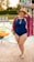 A person wearing a blue one-piece swimsuit and a wide-brimmed hat, standing by the edge of a swimming pool. They have a colorful, patterned cover-up draped over their shoulders and appear to be smiling joyfully. In the background, several inflatable rings float in the pool, and a waterslide is visible, suggesting a lively and fun summer setting.