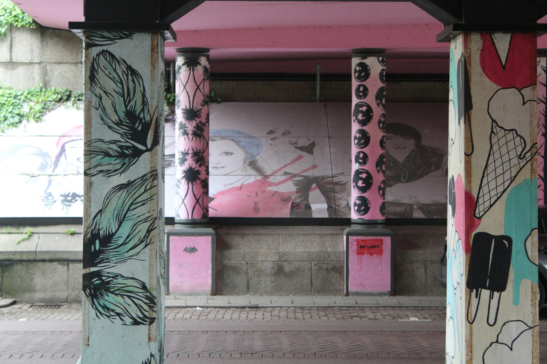 Painted columns with various artistic designs, including feathers and abstract faces. Murals in the background display stylized portraits with vibrant colors, adding an urban and creative atmosphere.