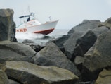 A rescue boat from a fire department is navigating through a rocky sea area with visible splashes of water. It is surrounded by large boulders along a coastline under a cloudy sky.